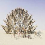 A group of four people with bicycles stand in front of a large, intricate wooden lotus flower sculpture on the flat, sandy desert at BurningMan. Other people and bikes appear in the distant background, capturing unique moments for BurningMan photos.