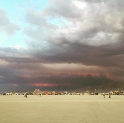 A wide, sandy desert with scattered people and vehicles in the distance under a dramatic sky. Thick, dark clouds with hints of red and purple loom overhead. Art structures evoke a festival, reminiscent of surreal BurningMan photos impossibles.