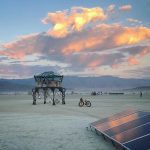 A futuristic metal structure stands on a barren desert plain at sunset, evoking surreal BurningMan images. Nearby, a bicycle and solar panels hint at off-grid life, while distant mountains and dramatic clouds enhance the remote atmosphere.