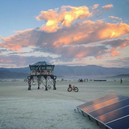 A futuristic metal structure stands on a barren desert plain at sunset, evoking surreal BurningMan images. Nearby, a bicycle and solar panels hint at off-grid life, while distant mountains and dramatic clouds enhance the remote atmosphere.