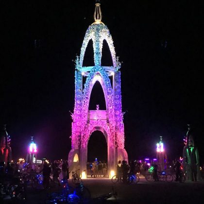 A tall, ornate archway glows with multicolored lights—pink, blue, and purple—against a dark night sky at BurningMan. Silhouetted people and bicycles gather around this striking structure, perfect for unforgettable BurningMan photos impossibles.