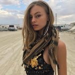 A woman stands on a sandy, open area at BurningMan with RVs and tents behind her. She wears a sequined black top, checked brown scarf, facial gems, long feather earrings, and loose wavy hair under the soft, cloudy light—perfect for photos impossibles.