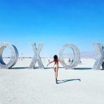 A woman in a bikini walks toward large metallic letters spelling “OXOX” in a desert landscape at BurningMan under a clear blue sky. A few people and bicycles are visible, creating the surreal vibe of classic BurningMan photos.