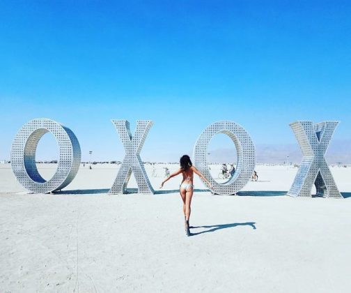 A woman in a bikini walks toward large metallic letters spelling “OXOX” in a desert landscape at BurningMan under a clear blue sky. A few people and bicycles are visible, creating the surreal vibe of classic BurningMan photos.