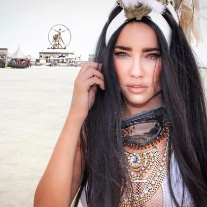 A woman with long black hair and intense blue eyes poses outdoors at Burning Man. She wears a feathered headpiece, layered jewelry, and intricate beading. In the background, people and a large circular art structure stand on the desert-like landscape.