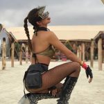 A woman with a long braided ponytail crouches outdoors on sandy ground, channeling BurningMan vibes in a gold top, black shorts, fishnet tights, platform boots, chunky accessories, and grey goggles. She sticks out her tongue for bold BurningMan photos.