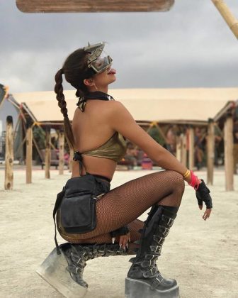 A woman with a long braided ponytail crouches outdoors on sandy ground, channeling BurningMan vibes in a gold top, black shorts, fishnet tights, platform boots, chunky accessories, and grey goggles. She sticks out her tongue for bold BurningMan photos.