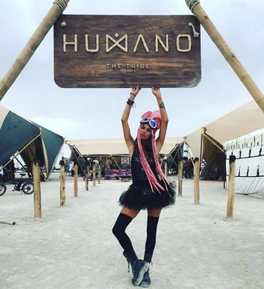 A woman with pink braids, goggles, black top, tutu skirt, and knee-high socks poses under a large wooden HUMANO THE TRIBE sign at an outdoor festival. This striking scene captures the photos impossibles spirit of Burning Man photos.