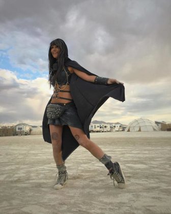 A person in a black hooded cape, chain top, studded skirt, and boots stands smiling on a sandy open area at Burning Man. They wear bracelets, a belt bag, and have thigh tattoos. Tents and domes dot the background beneath a cloudy sky.