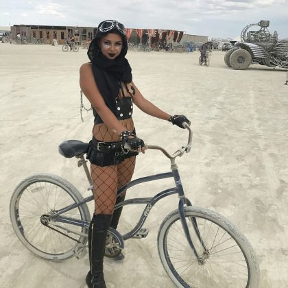A woman in black goggles, dark hood, crop top, shorts, fishnet tights, and boots stands on a dusty desert with her hands on a bicycle. Art cars and tents in the background suggest the iconic scene of BurningMan festival photos impossibles.