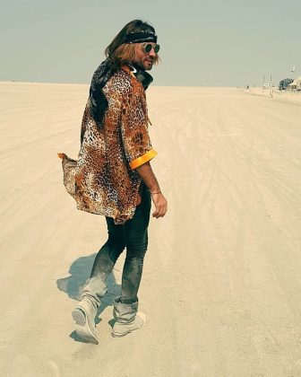 A person with long hair, wearing a leopard-print shirt, black pants, grey boots, and a black headband, walks on a wide, sandy landscape under a hazy sky—capturing that relaxed BurningMan vibe found in iconic BurningMan Photos.