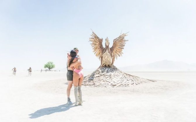 A couple embraces in the foreground on a sunlit, dusty plain at BurningMan. Behind them stands a large wooden phoenix sculpture with wings outstretched. A solitary tree and distant figures dot the pale desert landscape—true BurningMan Photos.