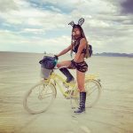 A woman in black festival attire, tall boots, and bunny ears stands with one foot on a yellow bicycle in the flat desert at BurningMan. She wears goggles and long braids; mountains and sparse figures dot the horizon in this photos impossibles scene.