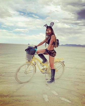 A woman in black festival attire, tall boots, and bunny ears stands with one foot on a yellow bicycle in the flat desert at BurningMan. She wears goggles and long braids; mountains and sparse figures dot the horizon in this photos impossibles scene.