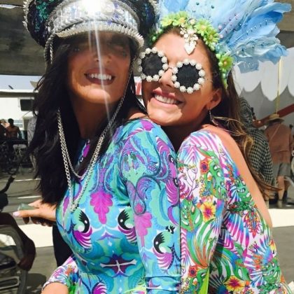 Two women smile and pose closely together at the BurningMan festival, wearing colorful, patterned bodysuits with My Little Pony and psychedelic designs. Both sport elaborate hats and sunglasses, including floral frames and a shiny sequined cap.