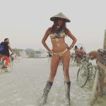 A woman in a gold bikini, tribal jewelry, and a conical hat stands confidently with hands on hips on dusty desert ground at BurningMan. Tall black boots complete her look amid bicyclists, tents, RVs, and art—capturing iconic Burning Man photos.