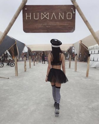 A woman in a black crop top, short skirt, police hat, and striped socks walks on dusty ground toward a “HUMANO THE TRIBE” sign at BurningMan, with tents, bikes, people, and art installations in the background.