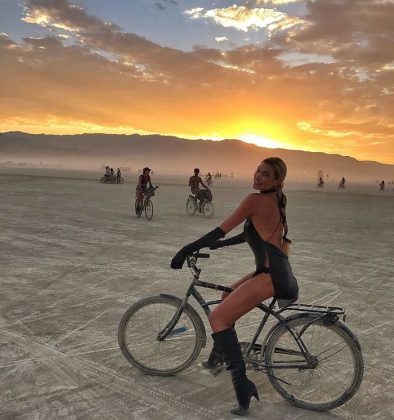 A woman in a black outfit, gloves, and boots sits on a bicycle on the flat desert at Burning Man sunset. Other cyclists trail behind as mountains are silhouetted against a dramatic sky—one of those photos impossibles to capture elsewhere.