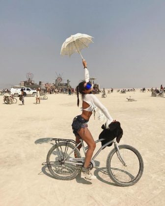 A woman poses with one leg up on a bicycle in the desert at Burning Man, holding a white parasol overhead. She wears a white top, blue shorts, boots, and blue goggles. Bikes, people, and art installations appear under a hazy sky—one of those photos impossibles.