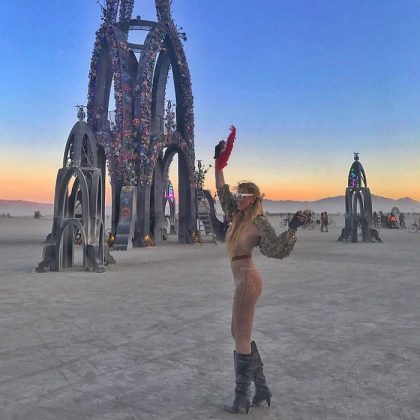 A woman in a sheer, sparkly outfit and boots stands on a desert playa at sunset, raising one arm and holding a red fan. Behind her are tall, ornate arch sculptures with flowers—an iconic scene from BurningMan photos.