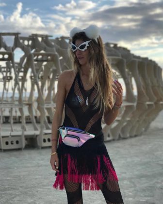 A woman stands outdoors at BurningMan, wearing a white fluffy cat ear headband, futuristic sunglasses, a black fishnet dress with pink fringe, and a shiny holographic fanny pack. Sculptural wooden art structures rise behind her under a partly cloudy sky.