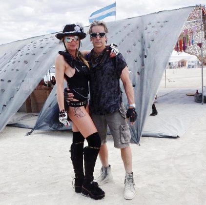A woman and man stand arm in arm on sandy ground at an outdoor event, capturing the spirit of BurningMan. She wears a black hat, sunglasses, bodysuit, gloves, and boots; he sports a gray shirt and shorts. Tents and flags fill the background—true Burning Man photos.