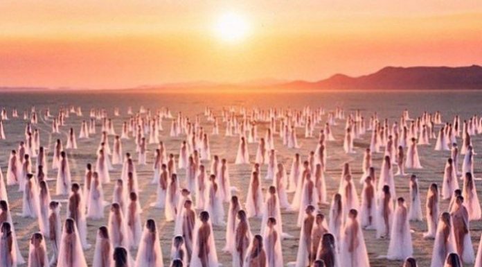 68 Photos Impossibles Du BurningMan Hundreds of women draped in sheer white veils stand spaced apart on a sandy, barren landscape at sunset. Reminiscent of BurningMan photos, the surreal scene glows with soft pink and gold light beneath an orange sky and distant mountains.