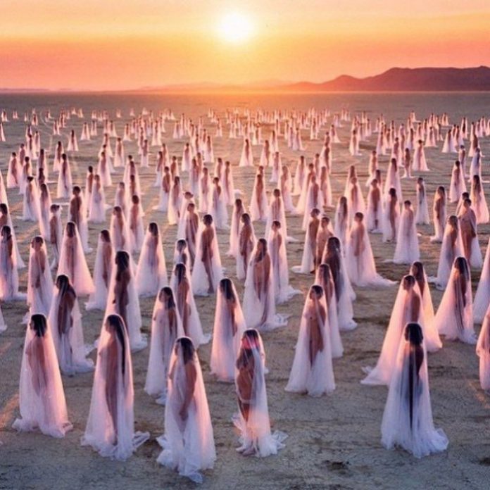 Des centaines de femmes drapées dans des voiles blancs transparents se tiennent à distance les unes des autres sur un paysage sablonneux et aride au coucher du soleil. Rappelant les photos de BurningMan, cette scène surréaliste s'illumine d'une douce lumière rose et dorée sous un ciel orange et des montagnes lointaines. 