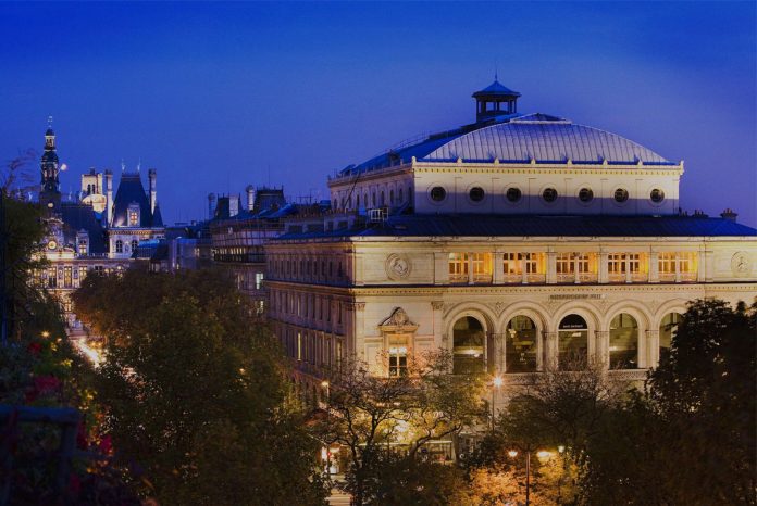 Un grand bâtiment historique - probablement l'Hôtel Britannique - avec de grandes fenêtres cintrées et des détails ornementaux est illuminé au crépuscule. Les lampadaires brillent en contrebas, les arbres bordent la rue et l'architecture classique apparaît dans le ciel bleu profond du soir. 
