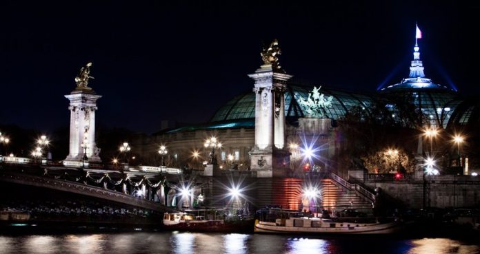 Vue nocturne du Pont Alexandre III de Paris avec ses lampadaires ornés et ses statues dorées. Derrière, le Grand Palais illuminé brille sous le drapeau français. Une vue parfaite à apprécier après vous être installé dans votre logement ou à l'hôtel Beaubourg à Paris. 