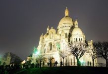 ÎLE DE FRANCE The illuminated Sacré-Cœur Basilica stands against a night sky, its white domes and ornate facade glowing. Silhouetted trees and a crowd of people are visible in the foreground on a grassy hill, with green lights casting accents on the scene.