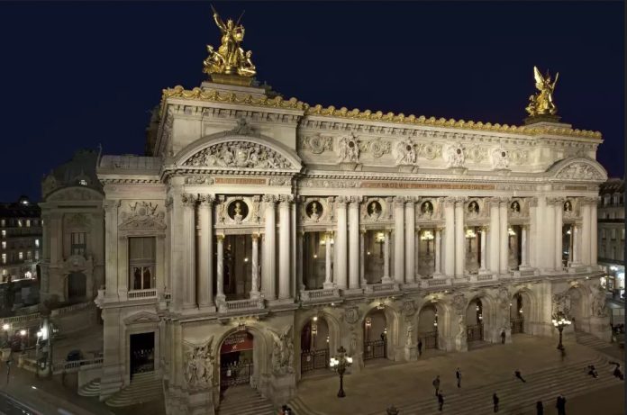 Un grand bâtiment orné, illuminé la nuit, avec de hautes colonnes, des fenêtres cintrées et des sculptures détaillées. Deux statues dorées se dressent sur le toit, et des lampadaires décoratifs illuminent les larges marches et la place devant l'édifice. 