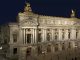 Opéra A grand, ornate building lit up at night, featuring tall columns, arched windows, and detailed sculptures. Two golden statues stand atop the roof, and ornate street lamps illuminate the wide steps and plaza in front.