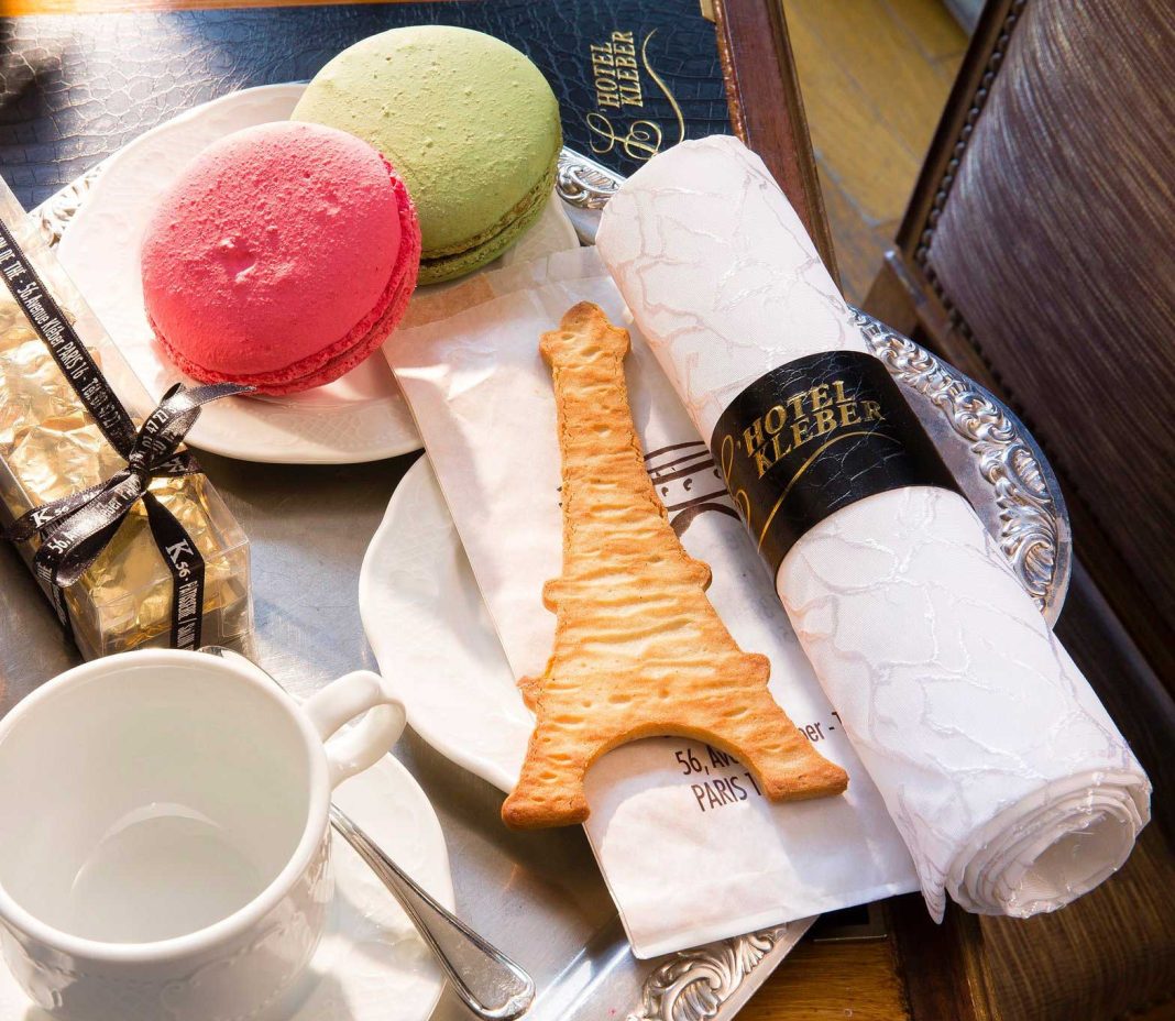 La table de l'Hôtel Kléber est décorée d'une serviette blanche entourée d'un anneau noir et or, de macarons pastel, d'un chocolat enrobé d'or, d'un biscuit Tour Eiffel et de la lumière du soleil, reflétant ainsi l'élégance des hôtels de luxe parisiens.