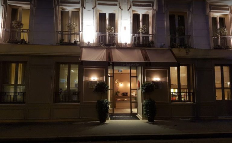 Vue nocturne de la rue de l'hôtel de charme "Le Tourville" à Paris. L'entrée est chaudement éclairée et flanquée de plantes en pot. Les fenêtres supérieures présentent de la verdure sur les balustrades, tandis qu'un éclairage doux met en valeur l'élégante façade de l'hôtel Le Tourville. 