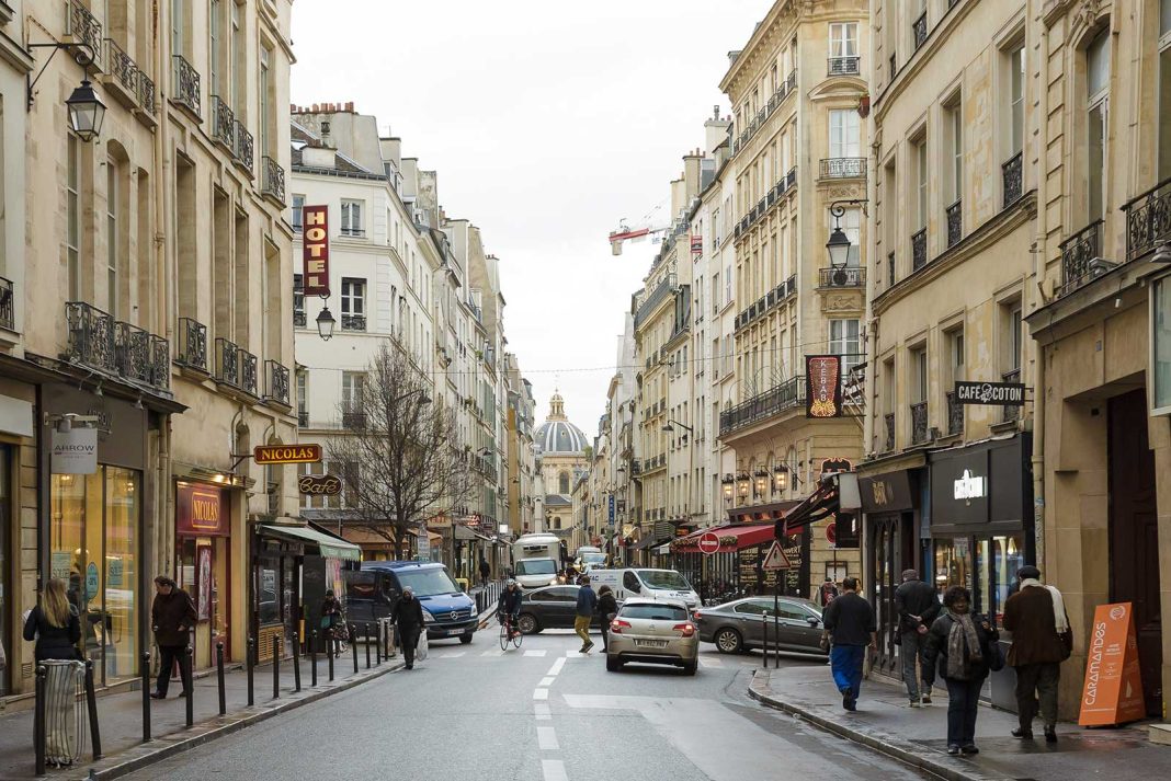 Une scène de rue animée à Saint-Germain, avec des voitures et des gens le long de grands immeubles beiges avec des balcons, des cafés, des magasins et un hôtel parisien. Les enseignes de l'Hôtel Rive Gauche et d'un bâtiment historique en forme de dôme s'élèvent au-dessus de la rue animée. 