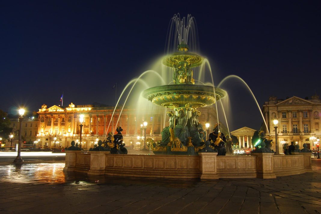 La nuit, une grande fontaine ornée de statues et de multiples jets d'eau est illuminée sur une grande place de la ville près de l'Hôtel Queen Mary. D'élégants bâtiments classiques brillent sous un ciel d'un bleu profond, avec des lampadaires et des reflets qui scintillent sur la chaussée humide. 