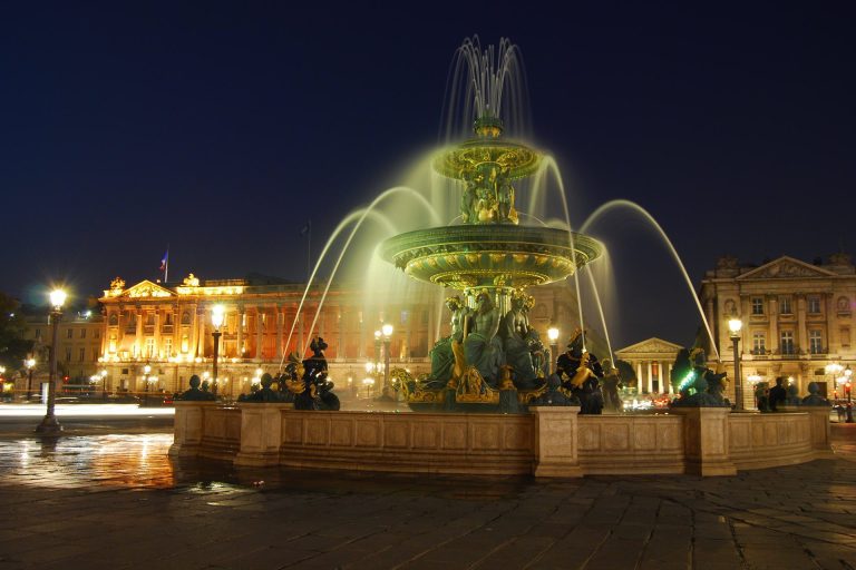 La nuit, une grande fontaine ornée de statues et de multiples jets d'eau est illuminée sur une grande place de la ville près de l'Hôtel Queen Mary. D'élégants bâtiments classiques brillent sous un ciel d'un bleu profond, avec des lampadaires et des reflets qui scintillent sur la chaussée humide. 