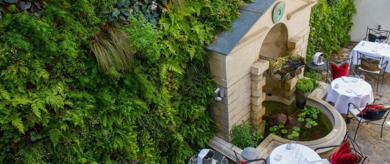Un jardin vertical luxuriant recouvre un mur à côté des tables de la salle à manger extérieure. Une fontaine en pierre avec des nénuphars et une petite statue est encadrée par de la verdure. Des nappes blanches, des verres et des chaises avec des coussins rouges et noirs créent une atmosphère de patio élégante et accueillante.  