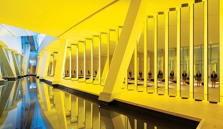 Un couloir moderne de la Fondation Louis Vuitton présente de hautes colonnes verticales éclairées en jaune qui se reflètent dans un bassin d'eau calme. Plusieurs femmes en tenues sombres sont assises sur des bancs, évoquant une installation artistique saisissante de répétition et de lumière. 