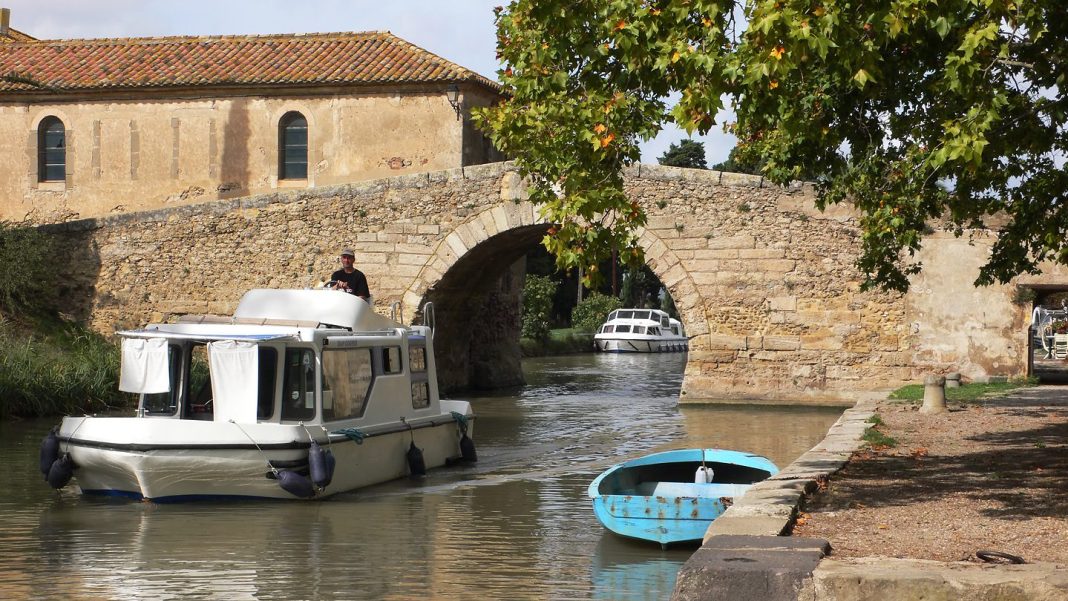 Bridge_over_Canal_du_Midi A small white houseboat and a blue rowboat float on a narrow canal beside an old stone arch bridge in France. A person stands on the houseboat as sunlight shines, with green trees and a rustic stone building in the background.