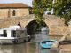Le long du canal… A small white houseboat and a blue rowboat float on a narrow canal beside an old stone arch bridge in France. A person stands on the houseboat as sunlight shines, with green trees and a rustic stone building in the background.