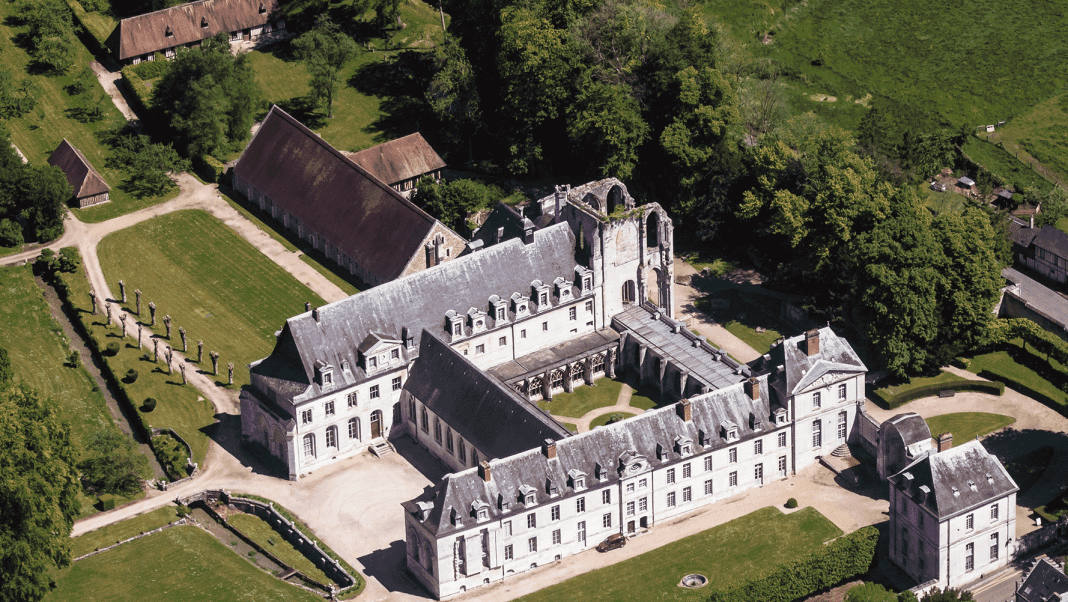abbaye-st-wandrille1 Aerial view of a large historic abbey along the route des abbayes in Haute-Normandie, with grey roofs and stone walls amid manicured lawns, tree-lined pathways, and woodland. The main buildings form a courtyard near outbuildings and green fields.