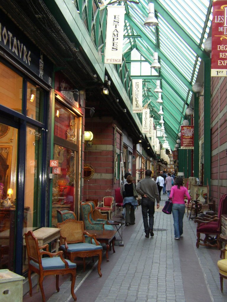 Ce marché aux puces de Paris est un couloir couvert avec un toit en verre. Il est bordé d'antiquaires qui exposent des chaises et des meubles anciens. Les acheteurs se promènent sous les enseignes tandis que la lumière du soleil passe à travers la lucarne à cadre vert. 