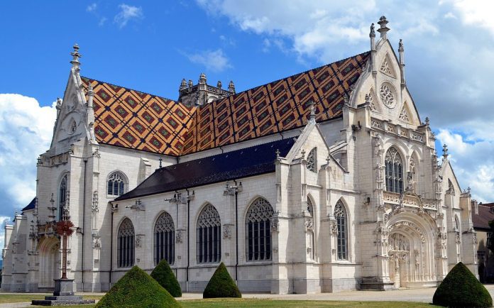 Une grande église de style gothique aux façades ornées de pierres blanches, d'arcs brisés et de vitraux constitue un véritable hymne de pierre. Son toit pentu est recouvert de tuiles colorées à motifs géométriques. Des buissons entretenus et un ciel bleu parsemé de nuages complètent la scène.  
