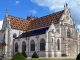 Un hymne de pierre A large Gothic-style church with ornate white stone facades, pointed arches, and stained-glass windows stands as a true hymne de pierre. Its steep roof features colorful, geometric patterned tiles. Manicured bushes and a blue sky with clouds complete the scene.