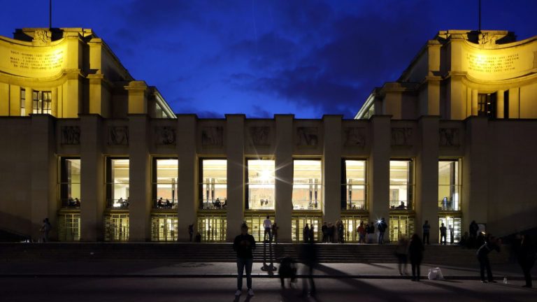 Vue nocturne du Palais de Chaillot à Paris, illuminé de lumières dorées sur un ciel d'un bleu profond. Des gens se tiennent debout et marchent sur les marches, leurs silhouettes étant encadrées par les grandes fenêtres lumineuses du Théâtre. 