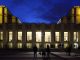 Le Théâtre à Paris A night view of the Palais de Chaillot in Paris, illuminated with golden lights against a deep blue sky. People stand and walk on the steps in front, their silhouettes framed by the Théâtre’s large, glowing windows.