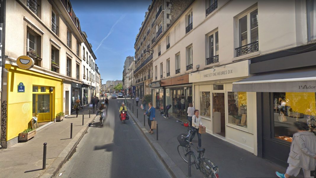 A narrow Parisian street lined with shops and cafes offers lively sunlit views, where pedestrians stroll and cyclists pass by. With balconies above, the scene invites you to enjoy un repas rapide or manger sur le pouce under a clear blue sky.