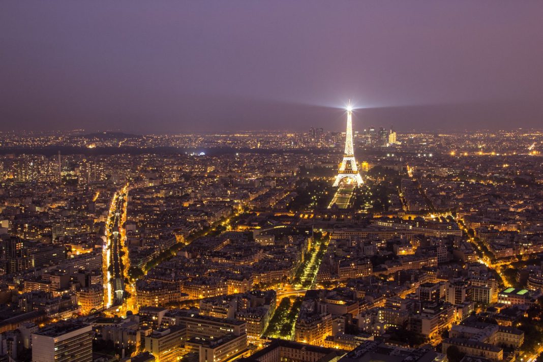 Une vue aérienne nocturne de Paris montre la ville illuminée par les lampadaires. La Tour Eiffel se dresse au centre de la scène, illuminée par les projecteurs, tandis que des monuments tels que l'Hôtel Renoir Paris se fondent dans les avenues animées qui s'étendent vers l'horizon brumeux. 